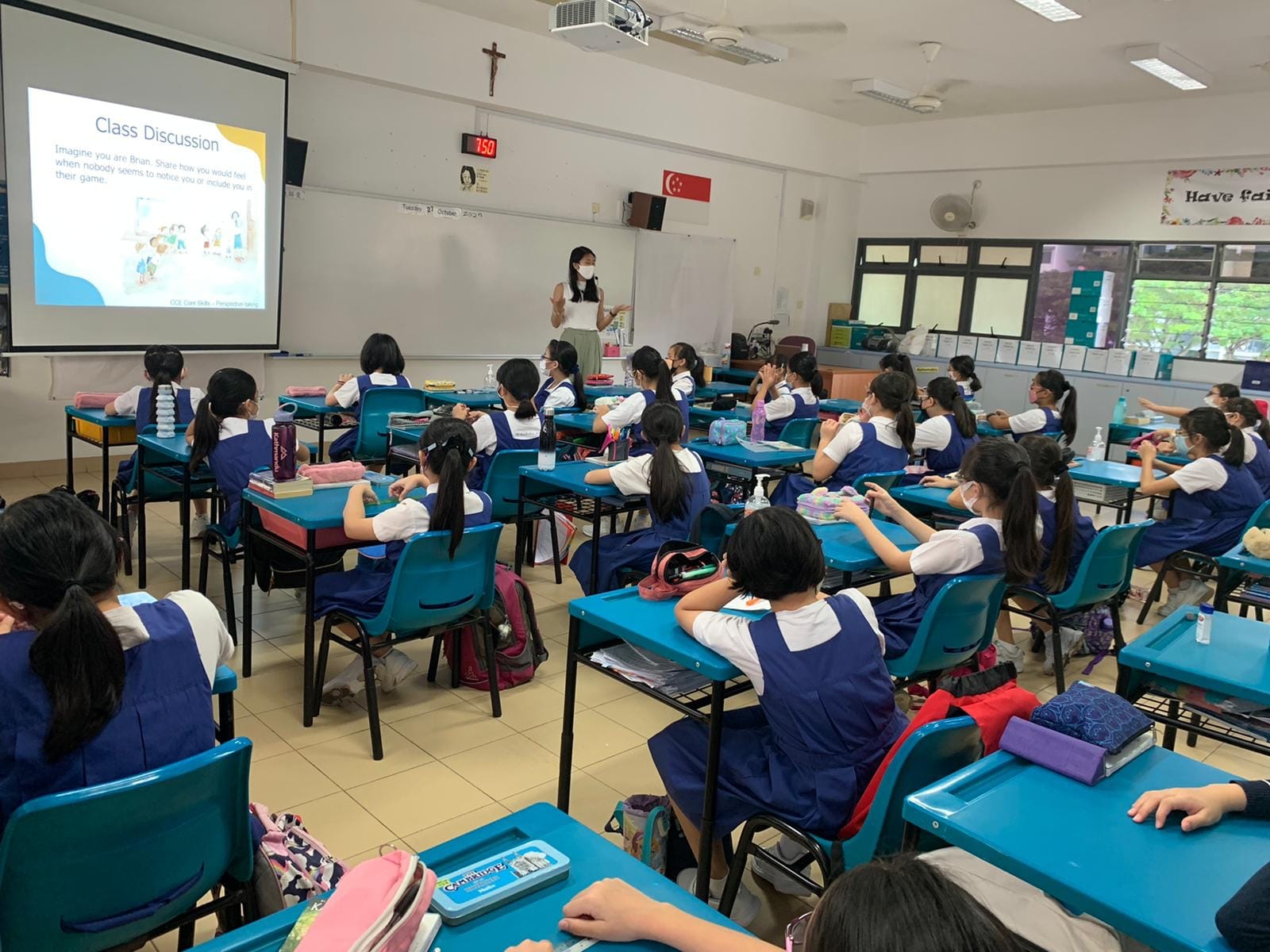 A teacher stands at the front of a classroom while girls in blue uniforms sit at teal desks facing a Class Discussion slideshow.